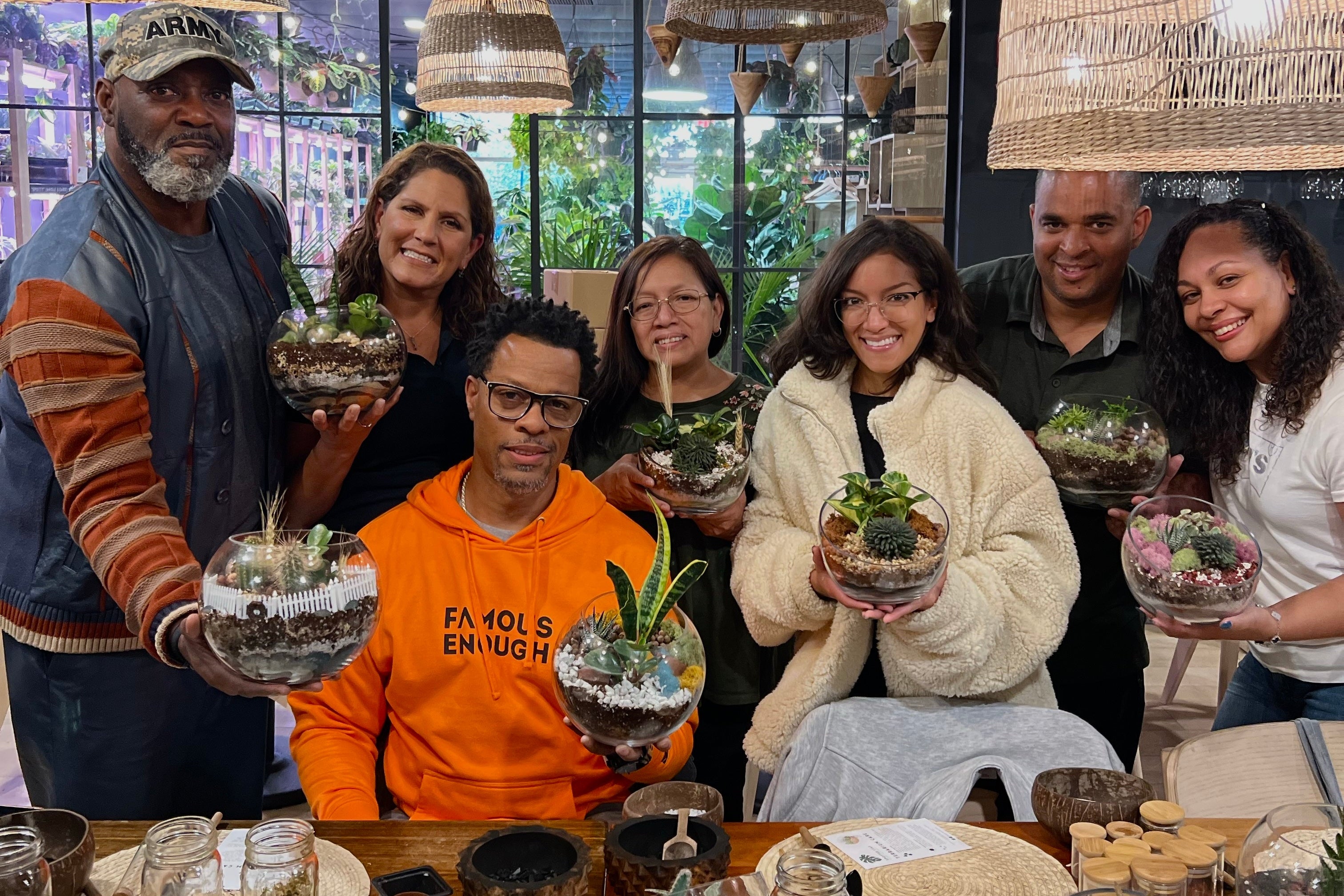 A group of seven people smiling and holding small glass terrariums with succulents in a bright indoor workshop or studio. The scene includes hanging woven pendant lights, large windows, and a wooden table with small pots and glass containers.