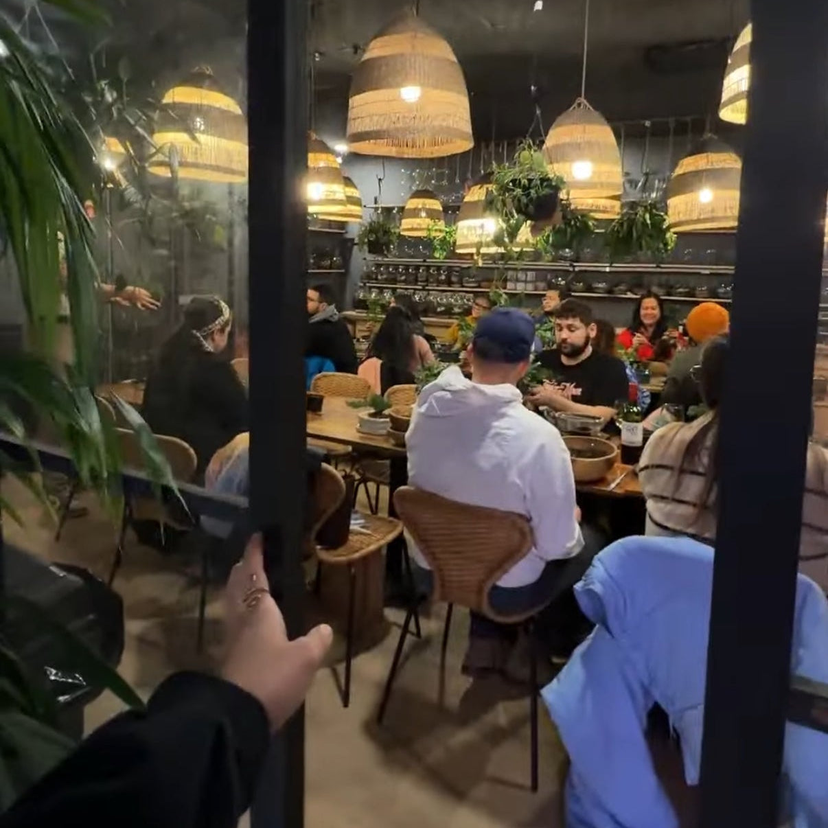 A view through a glass door into a cozy class with people seated at tables under warm hanging pendant lights. A hand is visible holding the glass door frame in the foreground.