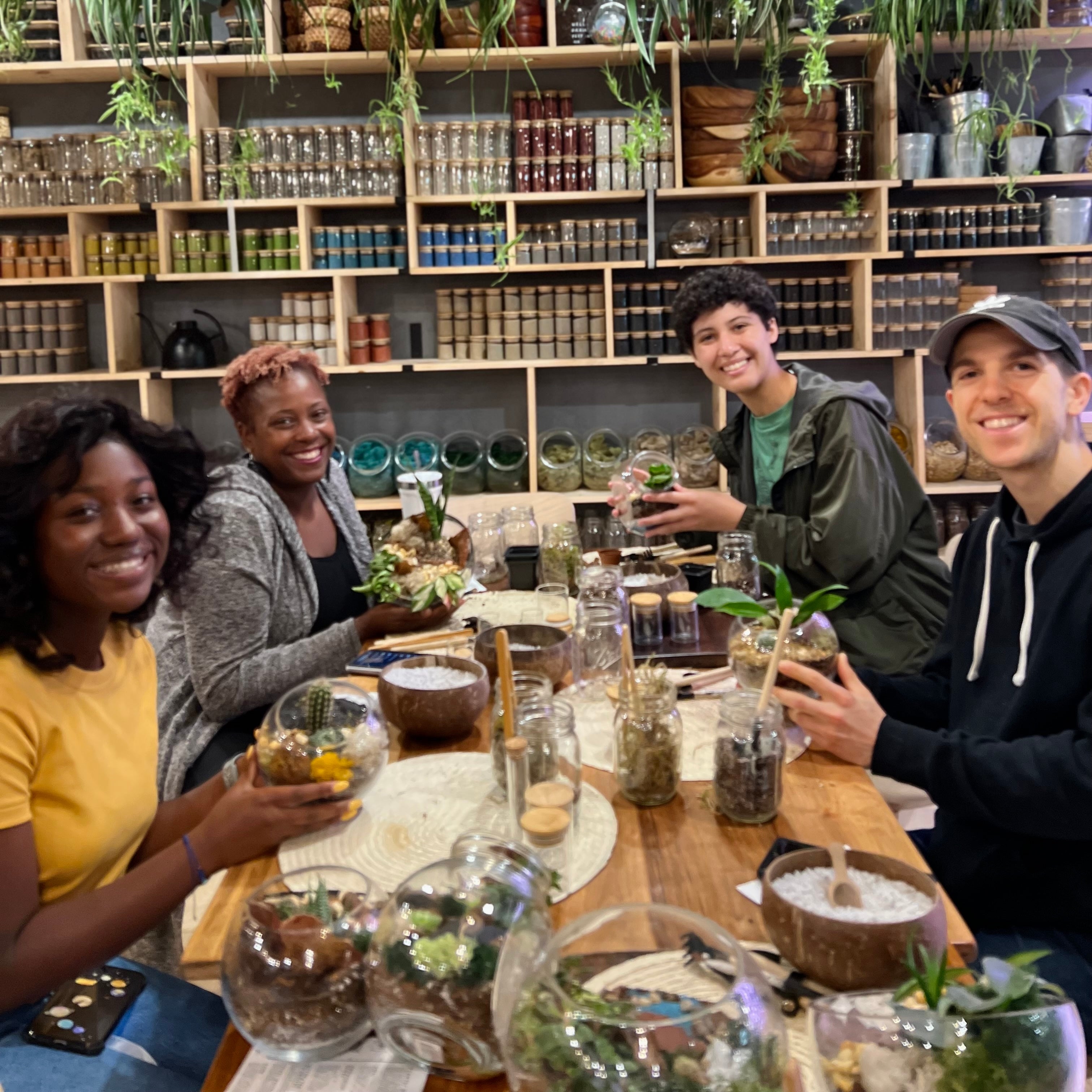 Four people smiling and posing together in a cozy, plant-filled craft studio, holding small terrariums and bowls filled with soil, rocks, and small green plants. The background features wooden shelves stocked with numerous glass jars and hanging greenery.