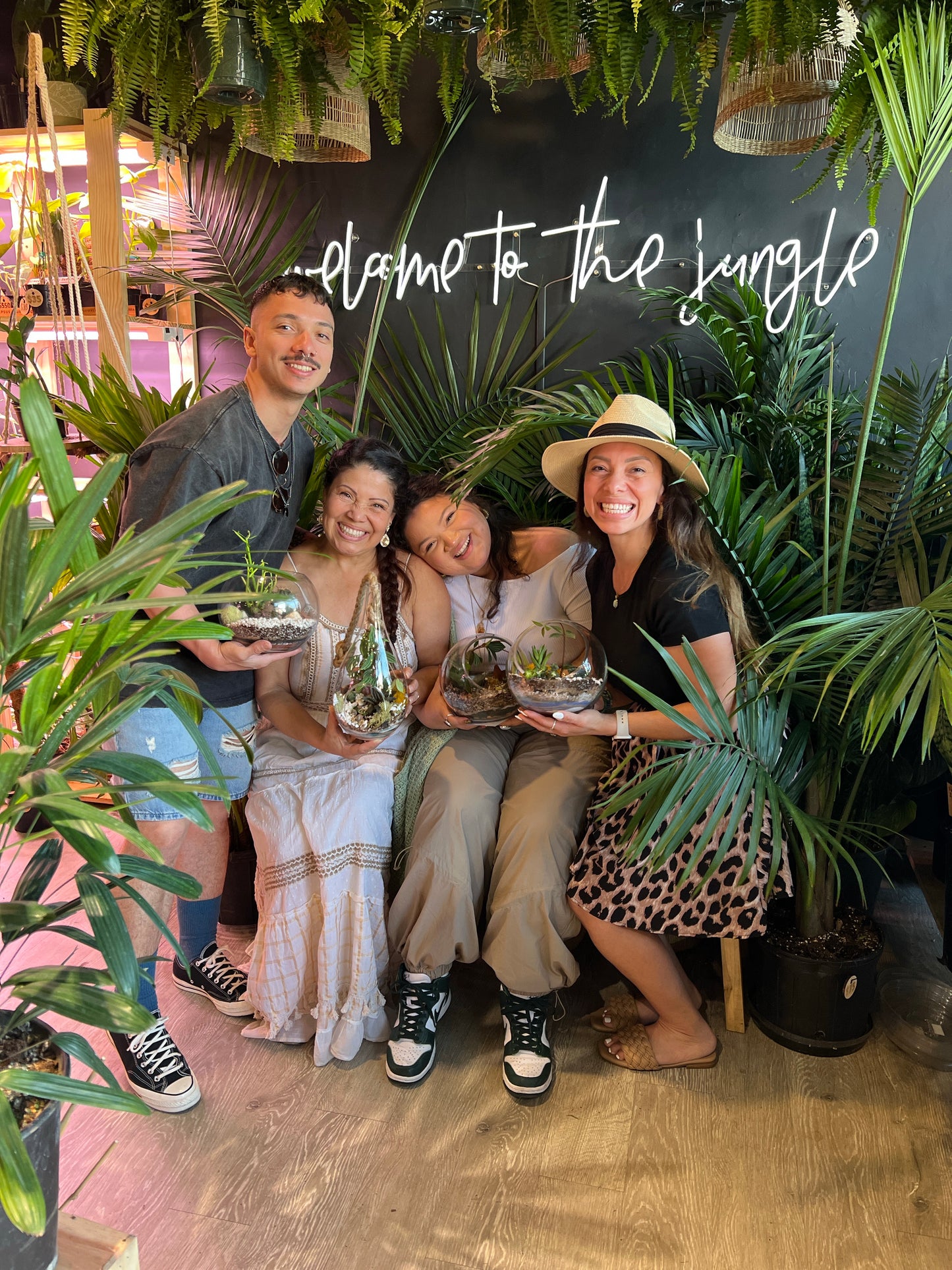 Four people posing together in a tropical, jungle-themed indoor setting with plants and a black wall reading 'welcome to the jungle'. Each person holds a small glass terrarium with green plants.