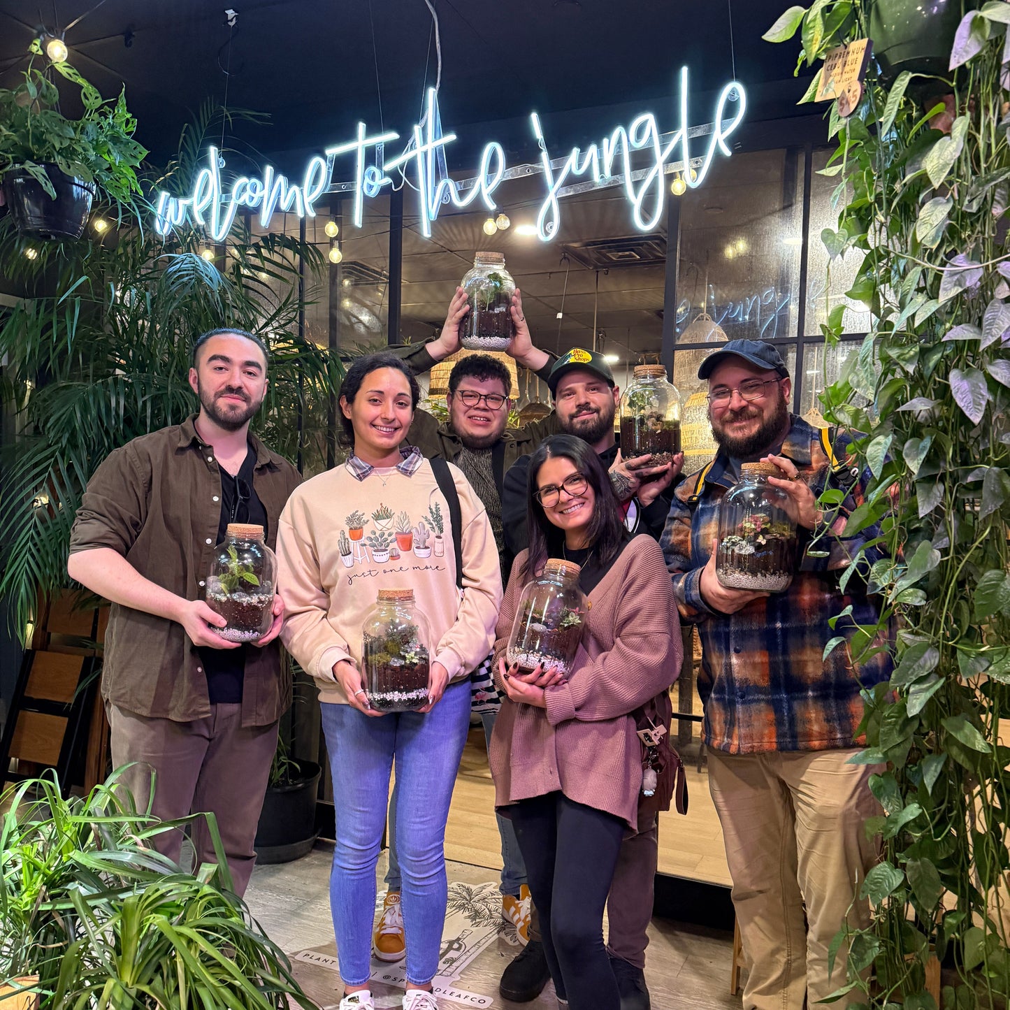 A group of six people standing together in a plant-filled indoor space with a glowing 'welcome to the jungle' sign, each holding a small glass terrarium with a plant.